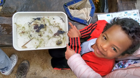 Nature school sessions in the woodland - A child smiles at the camera whilst pointing at a pond dipping sample in a white tray. 