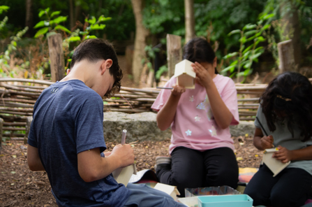 Three children sitting in the woodland area of Camley Street Natural Park decorating wooden bee hotels