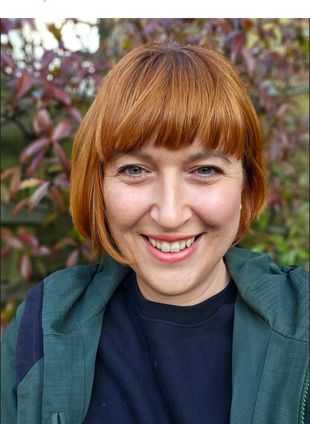 A woman with short red hair and a fringe, smiling at the camera. She is wearing a dark top and a green jacket, with a blurred background of green and reddish leaves