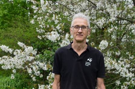 Mathew standing outside in front of a tree with white blossom. He is wearing a black t-shirt with the white London Wildlife Trust logo on it.
