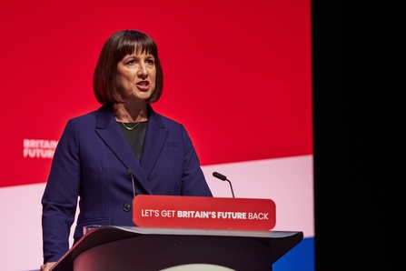 Chancellor Rachel Reeves stood at a podium giving a speech. A sign on the podium reads Let's get Britain's future back