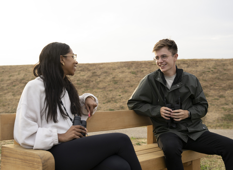 Two people sitting on a bench smiling and holding binoculars