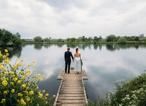 A couple, one in a wedding dress the other in a suit, stand on the edge of a bridge looking out at an expanse of water framed by flowered bushes